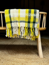A handwoven blanket with a geometric pattern in yellow, green, and ecru colors, displayed on a wooden chair.