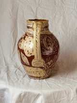 A vintage ceramic pitcher with a brown and beige floral pattern, featuring a tall handle and a small spout, set against a draped white fabric background.