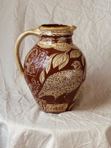 A brown and beige ceramic pitcher with a detailed bird and leaf pattern, set against a draped white fabric background.