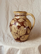 A vintage ceramic pitcher with a brown and beige floral pattern, featuring the text 'The Art of' on the front, set against a draped beige fabric backdrop.