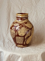 A decorative ceramic vase with a glossy dark brown and cream glaze, featuring hand-painted fruit and bird motifs, set against a wrinkled beige fabric backdrop.