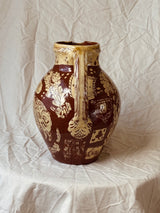A vintage ceramic pitcher with a rounded body, a vertical handle, and a flared rim, decorated with a glossy dark brown glaze and raised cream-colored floral and geometric patterns, set against a draped off-white fabric backdrop.