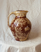 A brown and beige ceramic pitcher with hand-painted floral and landscape motifs, set against a draped cream fabric backdrop.