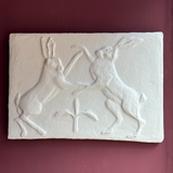 Relief sculpture of two hares on a rectangular stone block against a red background