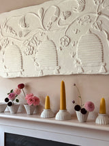 A decorative white plaster wall relief featuring stylized beehives and floral motifs, mounted above a white mantel decorated with yellow taper candles and small pink and burgundy flowers in white ribbed pots.