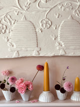 Decorative wall art featuring two stylized beehive motifs and floral vine details in an off-white textured finish, displayed above a shelf with pink flowers and yellow taper candles.