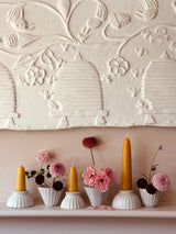 A styled interior scene showing a white mantel with several small white ribbed candle holders, yellow taper candles, pink flowers, and a large cream embossed wall panel above featuring birds and floral motifs.