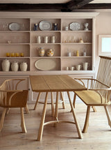A wooden trestle table made of oak, with a smooth finish and visible grain, accompanied by matching chairs. The table is set in a room with a wooden floor and a shelf of crockery in the background.