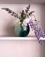 Vase with green and blue gradient holding purple flowers on a white surface.