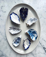 Seven blue and white patterned oyster shells arranged on an oval white ceramic platter over a light gray marble surface.