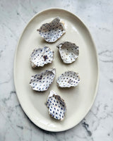 Six white oyster shells with blue polka dots arranged on an oval cream ceramic plate, set on a white and gray marble surface.