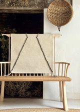 Wooden bench with a woven blanket draped over it against a white wall.