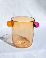 A translucent amber wine cooler, featuring a small orange handle on one side and a round magenta-pink accent on the other, set against a white fabric backdrop.