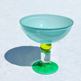 A translucent light blue-green ice cream coupe glass with a vibrant green circular base and a yellow accent at the stem, photographed on a white textured surface with a hard shadow.