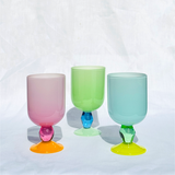 Three pastel-colored stemless goblets with faceted bases in pink, green, and light blue, arranged on a white fabric background.