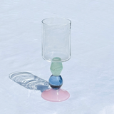 A clear glass goblet with a decorative stem featuring a light green faceted bead, a blue glass sphere, and a pink circular base, set on a white textured surface with a soft shadow.