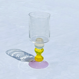 A clear glass with a cylindrical top and a sculptural base featuring stacked translucent yellow and light blue beads on a lavender-pink circular base, set on a white textured surface with a soft shadow.