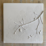 A square white ceramic tile featuring an embossed relief design of oak leaves and acorns, displayed on a rustic wooden surface.