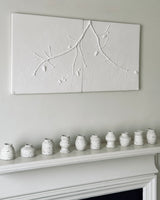 A two-panel white plaster wall art piece featuring a raised branch motif, displayed above a white fireplace mantel with a row of small speckled ceramic vases against a light gray wall.
