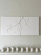 A two-panel white wall art piece featuring raised branch and leaf motifs, mounted on a light neutral wall above a shelf with small speckled ceramic vases.