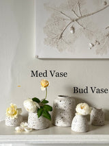 A set of speckled ceramic bud and medium flower vases displayed on a white mantelpiece against a light beige wall, with a framed botanical print above. Text overlays read 'Med Vase' and 'Bud Vase'.