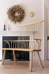 A high back settle bench made of oak and ash wood, hand-carved and finished with a tactile ripple design, positioned in a room with a white wall, wooden floor, a fireplace, and a decorative wreath on the wall.