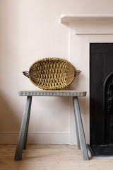 A woven oval basket with dark handles sits on a gray wooden stool in front of a cream wall and black fireplace.