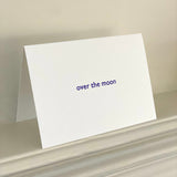 A white folded greeting card propped open on a light-colored surface, with the words 'over the moon' printed in small blue text on the front.