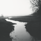 Black and white photo of a stream winding through grassy fields with trees on the right.