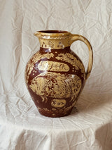 A vintage ceramic pitcher with a brown and gold hand-painted design featuring a sheep, the words 'Huff folk', and 'Bon', set against a draped off-white fabric background.