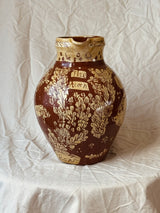 A brown and gold ceramic pitcher with a floral pattern and the words 'The Korn' on the front, set against a wrinkled off-white fabric backdrop.