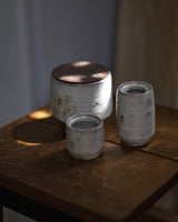 Three rustic handmade ceramic vessels with a textured off-white glaze and a dark metallic lid, displayed on a dark wooden table in warm, moody lighting.