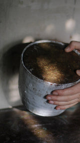 A close-up of a person's hands holding a large, rustic metal bucket filled with dark brown liquid, set against a dimly lit, textured gray background.