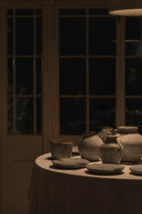 A group of rustic handmade ceramic vases and plates arranged on a draped tablecloth, set against a dark window at night with warm ambient lighting.