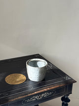 A light gray textured ceramic planter pot with a matching round brass tray, displayed on a dark wooden table against a plain beige wall.