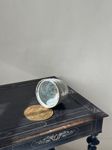 A distressed silver metal bucket lying on its side on a black wooden table, next to a round brass coin or medallion, set against a plain light gray wall.