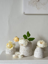 Three speckled ceramic vases in graduated sizes arranged on a white mantel against a neutral wall, styled with white and pale yellow peonies and green leaves.