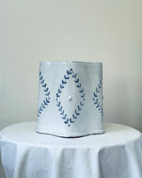 White lampshade with blue leaf pattern on a white tablecloth against a plain background