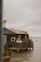 A rustic wooden fishing shack on a pier over murky water under an overcast sky, with lobster traps stacked on the deck and a life preserver hanging on the wall.