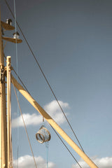 A close-up outdoor shot of a wooden mast and rigging against a blue sky with a few white clouds. A small coiled rope or sail component hangs near the center.