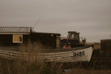 A weathered white wooden boat with the registration number JH85 painted on its side, resting in tall dry grass next to a dark wooden structure under an overcast, hazy sky.
