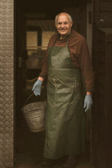 An older man wearing a green work apron, brown shirt, and blue gloves stands in a dark doorway holding a metal bucket.