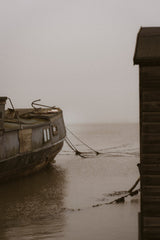 A moody, sepia-toned photograph of a rustic houseboat moored in calm, misty water next to a wooden dock or building, with a soft, overcast sky.