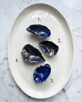Four small blue and gold painted oyster shells arranged on a white oval plate over a light marble surface, with numbered labels 1, 2, 3, and 4.