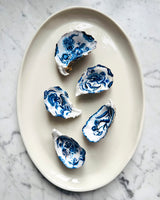 Five blue and white patterned oyster shell dishes arranged on an oval cream ceramic plate, set on a light gray marble surface.