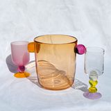An amber translucent bucket-style handblown glass wine champagny cooler with two handles, flanked by two small colorful stemmed glasses on a white fabric backdrop.