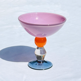 A decorative glass ice cream coupe with a wide, shallow pink bowl, a bright orange spherical stem accent, a clear faceted stem, and a blue-tinted circular base, photographed on a white textured surface.