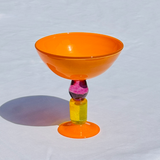 An orange translucent ice cream coupe glass with a purple and yellow marbled stem, standing on a white textured surface with a soft shadow.