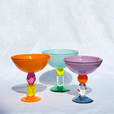 Three colorful glass ice cream coupes with wide bowls and decorative stems, shown on a white fabric background. The goblets are orange, light blue, and lavender purple, with multicolored glass accents in pink, yellow, green, blue, and clear.