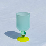 A translucent light blue goblet glass with a bright green stem and a vibrant yellow circular base, set against a white textured fabric background.